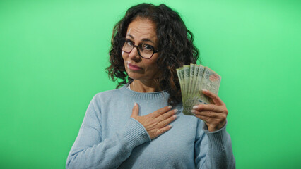 Middle-aged woman holding argentine pesos with hand on chest against green wall, expressing...