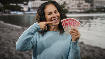 Woman smiling holding israeli shekels while gesturing okay on a seaside beach with blurred water...