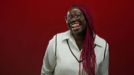 African american woman with glasses and long braids playfully sticking out her tongue against a vibrant red background, expressing joy and fun in a stylish outfit.