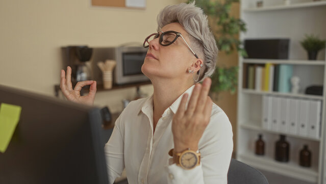 Woman wearing white shirt making ok sign with hands at office desk in front of computer monitor; mindfulness practice.