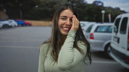 Fototapeta premium Young hispanic woman smiling covers eye with hand among parked cars on sunlit street outdoors; playfulness.