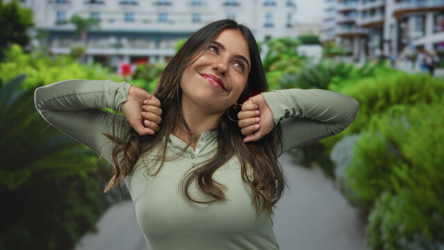 Young woman stretches arms beside lush green bushes on a sunlit paved path in front of building; serenity.