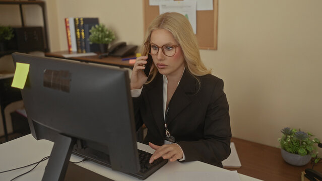 Woman talking on a phone in an office while reviewing documents, showcasing a productive work environment with a computer desk setup indoors and professional attire.