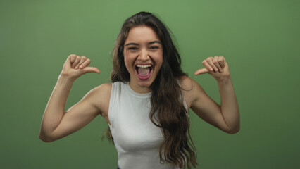 Hispanic young woman smiling broadly and pointing with thumbs to chest in a green studio; confidence.