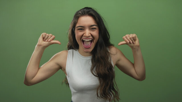 Hispanic young woman smiling broadly and pointing with thumbs to chest in a green studio; confidence.