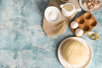 Plate with fresh raw dough, wheat, sugar, jug of milk and wooden holder of eggs on blue background
