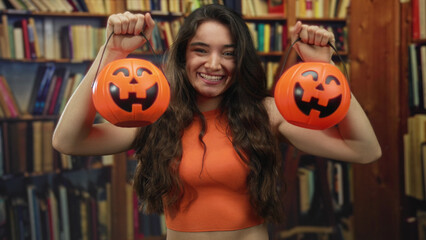 Young hispanic woman smiles while holding two orange pumpkin buckets in a library; halloween excitement.