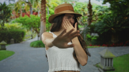 Obraz premium Hispanic teenage woman wearing straw hat and white tank top extends palm for a stop gesture in green park; rejection.