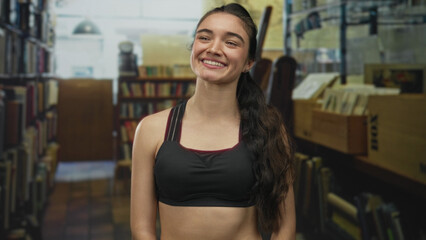 Young hispanic woman showing toned abs and smiling broadly in a library; confidence strength focus...