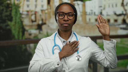 Woman wearing stethoscope stands solemnly with hand on heart and raised in oath gesture in front of...
