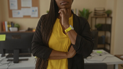 Woman standing thoughtfully in an office wearing a yellow shirt with braided hair, surrounded by...