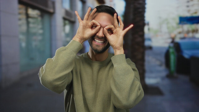 Hispanic man making glasses gesture with hands, smiling in an urban street setting, capturing a playful and humorous moment outdoors in a city environment.