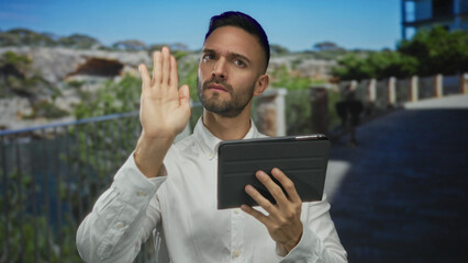 Fototapeta premium Hispanic man outdoors in urban setting using tablet with a focused expression, gesturing stop with hand, city backdrop featuring greenery and structures, conveying modern urban lifestyle.