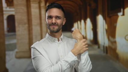 Young man holding wrist with expression of pain standing in a historic university campus surrounded by arches suggesting an academic setting.