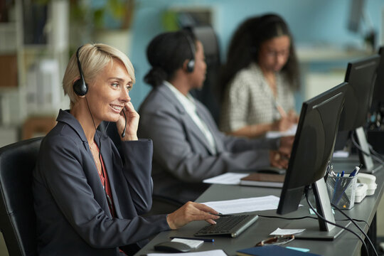 Caucasian middle aged woman wearing headset working at computer in modern office, sitting at desk with two colleagues in background using computers and headsets - Powered by Adobe