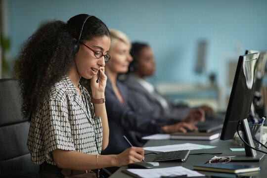 Young adult woman wearing headset talking on phone and taking notes while working at computer in modern office with two female colleagues in background