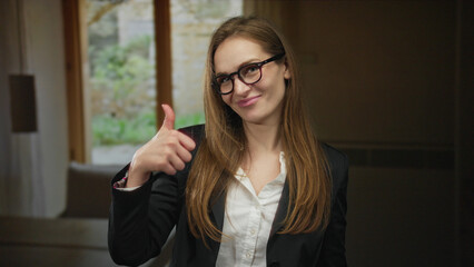 Woman wearing glasses and blazer showing a thumbs up gesture in a living room by a window;...