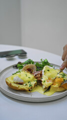 Person cutting tasty eggs benedict with hollandaise sauce on a cafeteria table one beside a smartphone capturing the essence of a casual breakfast moment.