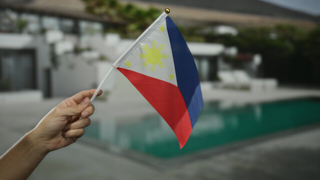Hand holding philippines flag by outdoor hotel pool, capturing a tropical and patriotic moment at resort setting under clear skies.