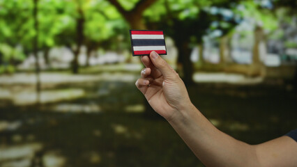 Man holding thai flag patch outdoors in park with green trees, showing hand against blurred natural background, showcasing craftsmanship and patriotism in bright daylight.