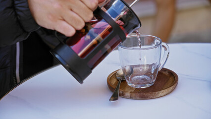 Hand pouring hot beverage from glass teapot into cup on table in cozy cafe setting, showcasing relaxation and warmth.