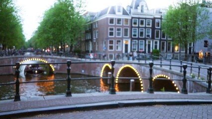 Blurred view of a lit bridge over a canal in amsterdam with historic buildings and green trees, creating a picturesque dutch cityscape.