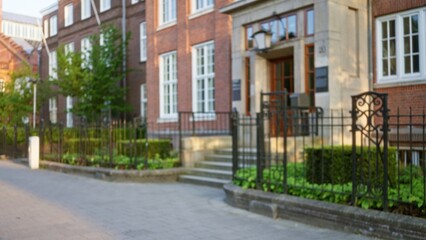 Blurred view of an elegant european city street with historic architecture, featuring defocused brick buildings, fences, and lush greenery under a soft daylight bokeh effect.