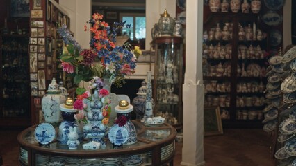 Blurred antique store with defocused porcelain vases and colorful flowers displayed in glass cabinet, surrounded by intricate dishes and art pieces creating a classic elegant atmosphere.