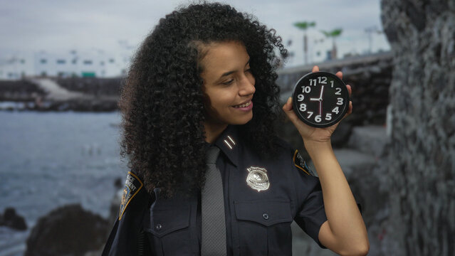 Woman in police uniform smiles holding clock by seaside, showcasing time and law enforcement in a beach setting with tropical vibes, emphasizing public service and punctuality.