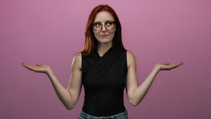 Confident woman with red hair wearing glasses stands against an isolated pink background with arms outstretched, exuding beauty and elegance in a stylish black top.