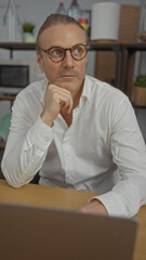 Fototapeta premium Mature man in an office setting wearing glasses and a white shirt, sitting thoughtfully at a wooden desk with modern decor and indoor lighting, suggesting a focused and professional atmosphere.