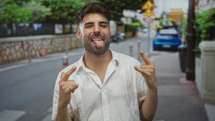 Man grinning and showing tongue while making rock horns gesture with both hands on busy street;...