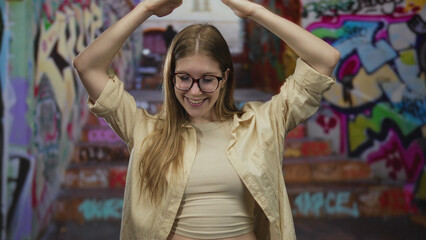 Woman raises arms overhead in graffiti street with vibrant graffiti walls and colorful tags; confidence.