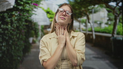 Fototapeta premium Young blonde woman in beige shirt touching neck near stone building with glasses reflecting sunlight; stress anxiety.
