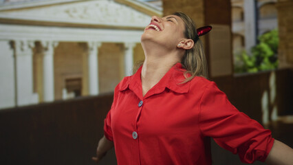 Woman in a red blouse tilts head back and spreads arms on an old town street beside a building;...