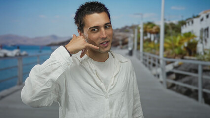 Fototapeta premium Young hispanic man standing on a seaside promenade gesturing a phone call against a coastal backdrop under sunny skies.