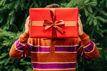 Person in cozy sweater holding large wrapped gift in front of face outdoors