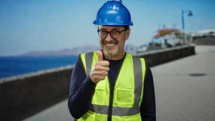 Hispanic man wearing a hardhat and safety vest giving a thumbs up on a seaside promenade under clear sky.
