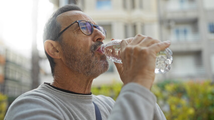 Mature man drinking water outdoors in urban park setting wearing glasses and gray shirt against city backdrop on a sunny day