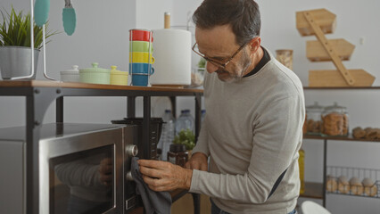 Fototapeta premium Hispanic man cleaning microwave in modern office kitchen, surrounded by colorful kitchenware and indoor plants, conveying a tidy, organized workspace atmosphere.