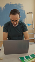 Middle-aged hispanic man using laptop at home with painting supplies in room showing home improvement setting.