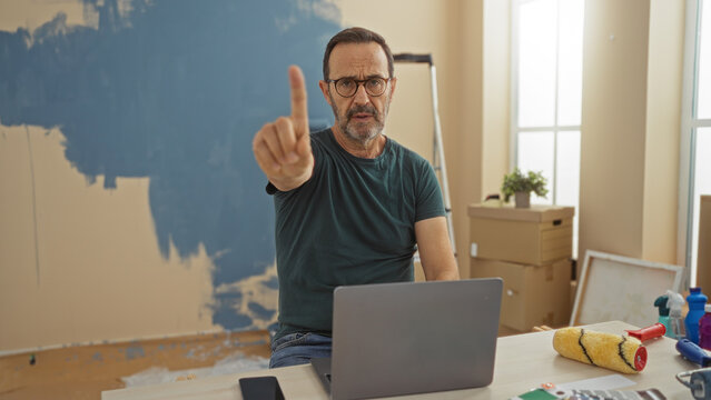 Hispanic man in glasses pointing finger while seated at desk with laptop in new home living room showing boxes and painting supplies. - Powered by Adobe