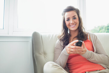 Woman holding smartphone while sitting on beige sofa by large window in living room, copy space