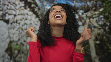 Woman clenches fists while smiling broadly with bright eyes in lush forest; revelation triumph...