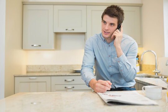 Man using smartphone while reviewing printed pages on kitchen island with coffee mug, copy space