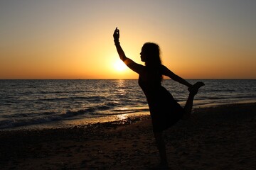 Fototapeta premium Silhouette of a woman balancing in a graceful pose on the beach at sunset, with the sun low over the sea and warm golden light illuminating the horizon and creating a peaceful coastal atmosphere.