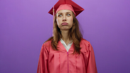Woman wearing graduation cap and gown looks upward against an isolated purple background, conveying...