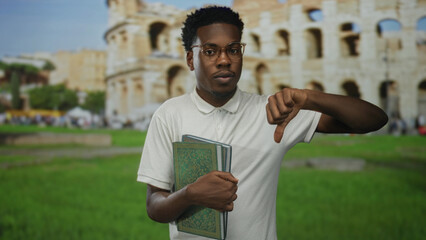 Young african american man wearing glasses holding green book shows thumb down in roman coliseum...