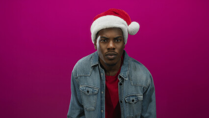 Man wearing santa hat and denim jacket stands in pink studio setting with direct gaze and visible face; holiday joy.