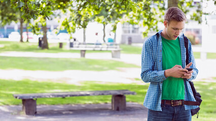 Fototapeta premium Man standing under leafy tree shade in urban park, checking smartphone beside wooden bench